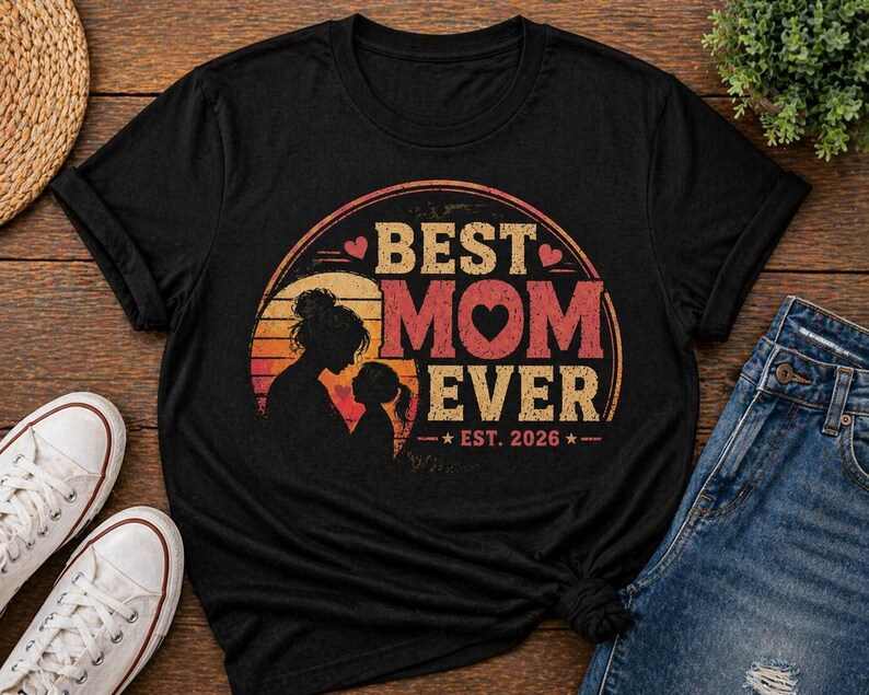 K&ouml;nnte beinhalten: Schwarzes T-Shirt mit Retro-Design, das "BEST MOM EVER" in orangefarbenen und roten Buchstaben zeigt. Das Design beinhaltet eine Mutter-Kind-Silhouette, mit dem Jahr "EST. 2026" darunter.