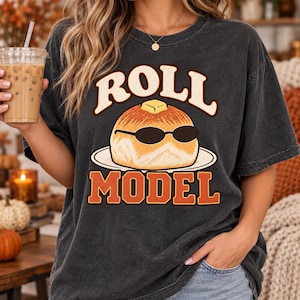 Könnte beinhalten: Ein anthrazitfarbenes Kurzarm-T-Shirt mit einer Grafik eines Brötchens, das eine schwarze Sonnenbrille trägt, auf einem Teller mit einem Stück Butter darauf. Der Text ROLL steht darüber und MODEL darunter in orangefarbenen Buchstaben.