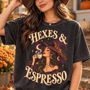 Può includere: Una t-shirt grigio antracite a maniche corte con una stampa grafica di una persona che indossa un cappello da strega viola con sopra un gatto nero, mentre tiene in mano una tazza di caffè fumante. Il testo recita HEXES & ESPRESSO in color crema.