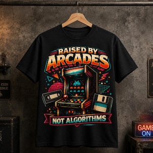 May include: A black short-sleeve t-shirt featuring a colorful graphic print of a vintage arcade cabinet, a VHS tape, and a floppy disk. The text reads RAISED BY ARCADES NOT ALGORITHMS in bold, stylized lettering with retro-inspired geometric shapes.