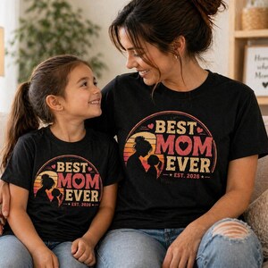 K&ouml;nnte beinhalten: Zwei schwarze T-Shirts mit dem Text "BEST MOM EVER" in einem kreisf&ouml;rmigen Design mit einer Mutter-Kind-Silhouette. Die Shirts haben auch den Text "EST. 2026". Mutter und Tochter tragen die passenden Oberteile.