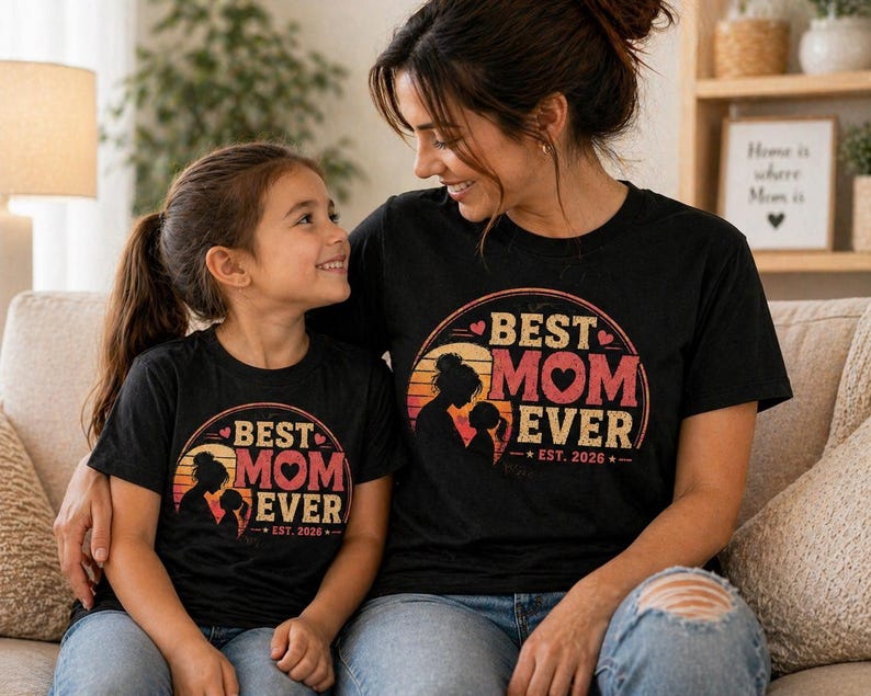 K&ouml;nnte beinhalten: Zwei schwarze T-Shirts mit dem Text "BEST MOM EVER" in einem kreisf&ouml;rmigen Design mit einer Mutter-Kind-Silhouette. Die Shirts haben auch den Text "EST. 2026". Mutter und Tochter tragen die passenden Oberteile.