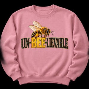 Könnte beinhalten: Ein altrosa Sweatshirt mit Rundhalsausschnitt und einer Grafik einer Honigbiene über dem Schriftzug UN-BEE-LIEVABLE. Die Biene ist in realistischen Gelb-, Schwarz- und Brauntönen gehalten, während die Schrift einen abgenutzten schwarz-gelben Look aufweist.