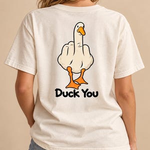 Puede incluir: Una camiseta de manga corta color hueso con un gráfico en la espalda de un pato blanco con patas y pico naranjas, mostrando el dedo corazón. Debajo del pato, el texto Duck You está impreso en letras negras en negrita.