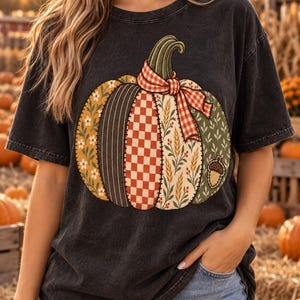 以下が含まれることがあります： チャコールグレーの半袖Tシャツ。花柄、ストライプ、チェック、麦の穂など、様々な布地風のパッチワークで構成された大きなカボチャのグラフィックが描かれており、赤と白のギンガムチェックのリボンとどんぐりのアクセントが添えられています。