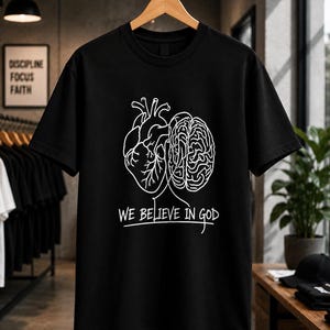 Peut inclure: Un t-shirt noir à manches courtes orné d'un graphisme en lignes blanches représentant un cœur humain et un cerveau côte à côte. Sous ces illustrations anatomiques, le texte "WE BELIEVE IN GOD" est imprimé en blanc avec un style manuscrit.