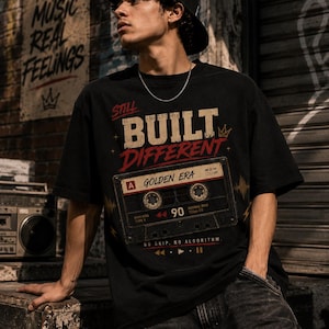 May include: Black graphic t-shirt with the text "STILL BUILT DIFFERENT" and a cassette tape design. The shirt features the words "GOLDEN ERA" and "90". The person is wearing a black baseball cap and a silver chain.