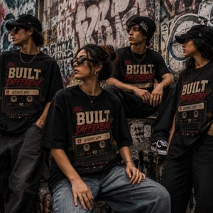 May include: Four people wearing black t-shirts with the text "BUILT DIFFERENT" and a cassette tape graphic. The shirts are paired with jeans and accessories like hats and sunglasses. The background features graffiti art.
