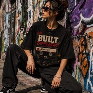May include: Black t-shirt with the text "Still Built Different" and a cassette tape graphic. The shirt is paired with black jeans and sneakers. The background features a colorful graffiti wall.