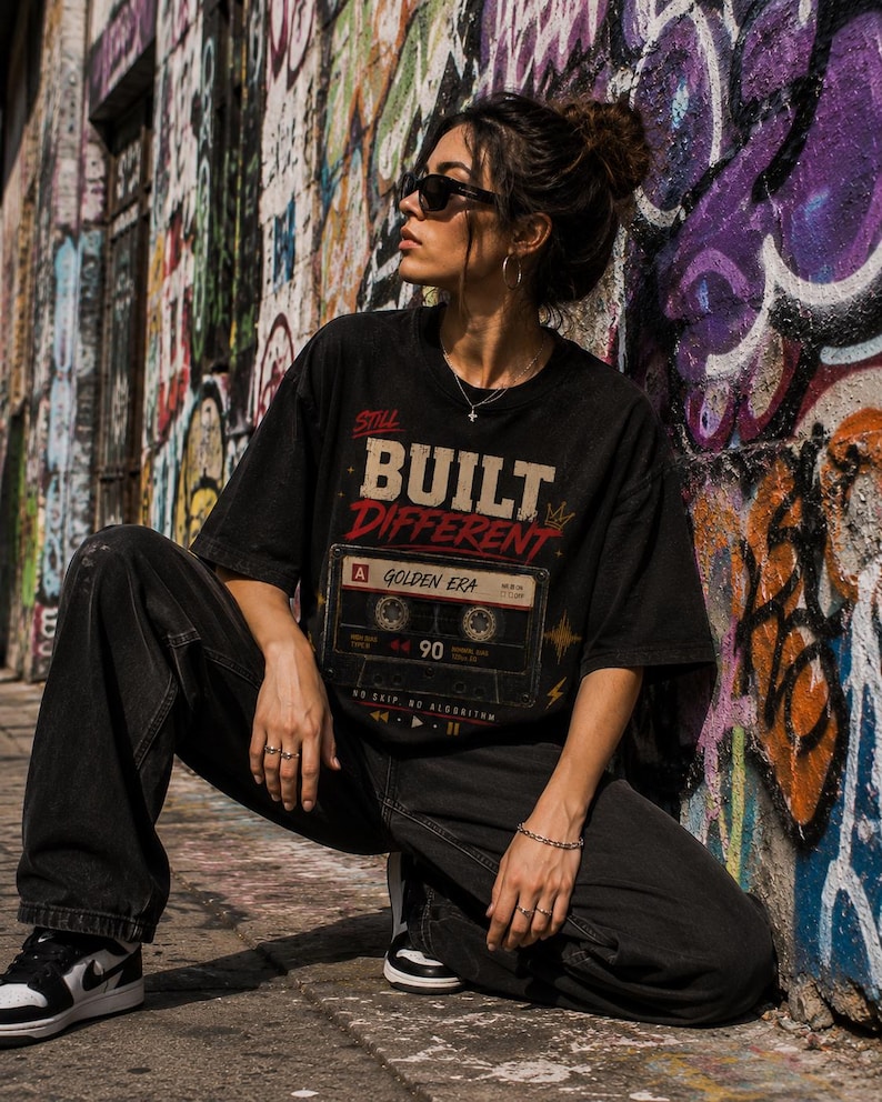 May include: Black t-shirt with the text "Still Built Different" and a cassette tape graphic. The shirt is paired with black jeans and sneakers. The background features a colorful graffiti wall.