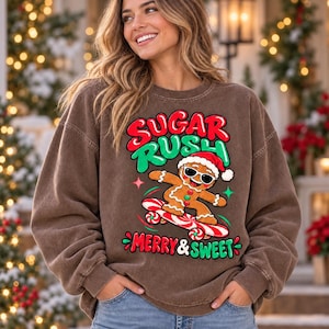 May include: A brown crewneck sweatshirt featuring a graphic of a gingerbread person wearing sunglasses and a Santa hat while riding a candy cane skateboard. The text reads SUGAR RUSH in red and green, and MERRY & SWEET in red and green.