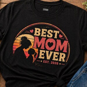 K&ouml;nnte beinhalten: Schwarzes T-Shirt mit einem kreisf&ouml;rmigen Aufdruck &bdquo;BEST MOM EVER&ldquo; in Rot und Beige, mit einer Silhouette von Mutter und Kind. Der Text &bdquo;EST. 2026&ldquo; steht darunter. Das Shirt wird mit blauen Jeans und wei&szlig;en Turnschuhen kombiniert.