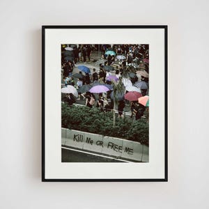 May include: A framed photograph shows a street scene with a crowd of people using umbrellas of different colours. The image includes the text "Kill Me or Free Me" on a concrete barrier.