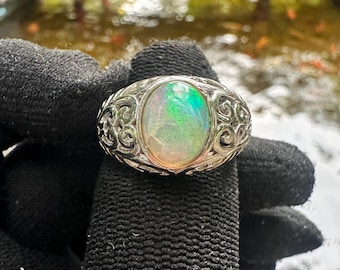 May include: A silver ring with an oval opal gemstone. The opal displays a play of color with green and orange hues. The ring has an ornate, filigree design around the gemstone. The ring is held up by a black glove.