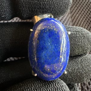 Puede incluir: Colgante ovalado de lapislázuli azul intenso con motas doradas, montado en una fianza plateada. La joya se presenta sobre un fondo negro texturizado.