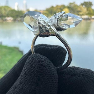 Peut inclure: Une bague en argent ornée de deux pierres de cristal de quartz claires et facettées. La bague a un anneau en argent poli et les cristaux sont montés sur un support en argent texturé. En arrière-plan, un lac et des arbres.