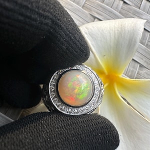 Könnte beinhalten: Ein silberner Ring mit einem großen, runden Opal-Edelstein. Der Opal zeigt eine Mischung aus Farben, darunter Orange, Grün und Rot. Der Ring ist von einem Kranz aus kleinen, klaren Edelsteinen umgeben. Der Ring liegt auf einer weißen Blume.