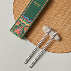 Puede incluir: Un par de palillos chinos plateados sobre un reposapalillos gris. En el fondo, una caja verde con diseños florales y de olas coloridas. La caja tiene la palabra "Bapmoo" impresa.