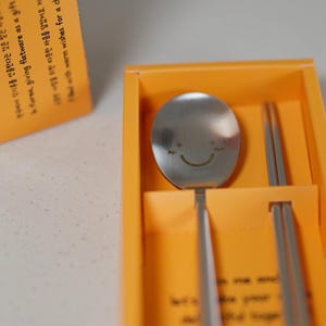Puede incluir: Una cuchara de acero inoxidable con una cara sonriente grabada en el cuenco, presentada con palillos a juego en una caja naranja. El juego está diseñado para regalar e incluye texto en coreano.