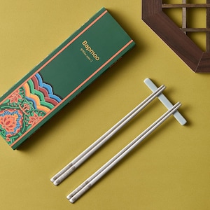 May include: Silver chopsticks and a rest are displayed next to a green box with the word "Bapmoo" and floral patterns. The chopsticks are on a yellow-green surface. The box has a decorative orange border.