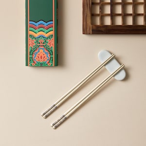 May include: A pair of light-colored chopsticks with decorative silver tips, resting on a pale gray chopstick rest. A green box with a colorful floral design is visible in the background, along with a wooden window.