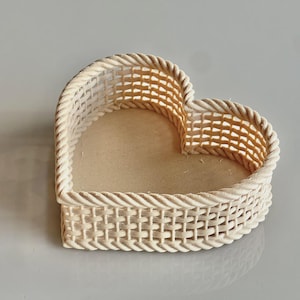 Op de afbeelding: Een hartvormige geweven mand in een natuurlijke crèmekleur. De mand heeft een geweven ontwerp met een touwachtige rand rond de randen. De binnenkant is lichtbeige, wat een charmant decoratief stuk creëert.