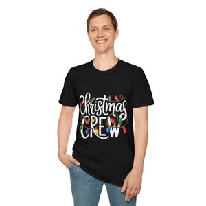 Könnte beinhalten: Schwarzes T-Shirt mit Rundhalsausschnitt und dem Schriftzug "Christmas CREW" in weißer Schrift sowie einer Lichterkette mit bunten Weihnachtslichtern. Das T-Shirt wird von einer Person in blauen Jeans getragen.