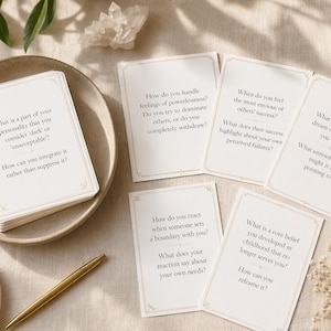 Pode incluir: Uma pilha de cartões brancos com bordas douradas e texto, juntamente com outros cartões, uma caneta dourada e uma tigela pequena com um colar dourado. Os cartões contêm perguntas sobre autorreflexão. Os itens estão dispostos em uma superfície bege.