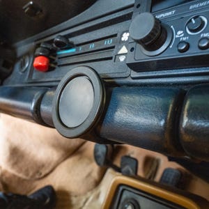 Puede incluir: Primer plano del interior del salpicadero de un coche. El salpicadero es negro con un soporte magnético redondo y negro para el teléfono. La radio tiene botones y un dial. Se ve un botón rojo. El interior es de color beige.