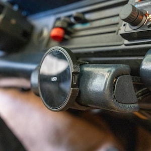 Puede incluir: Un soporte magnético negro para teléfono móvil fijado al salpicadero de un coche. El soporte tiene un diseño circular con una superficie negra y un borde plateado.