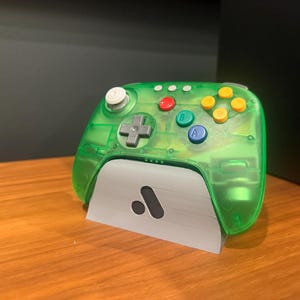 Könnte beinhalten: Ein transparent grüner Videospiel-Controller mit roten, blauen und gelben Knöpfen. Der Controller ruht auf einem weißen Ständer mit einem schwarzen Logo. Der Controller hat ein Steuerkreuz und einen weißen Joystick.