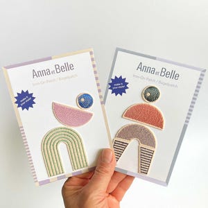 Könnte beinhalten: Zwei Anna et Belle Bügelpatches auf Präsentationskarten. Der linke Patch zeigt einen grünen Bogen, einen rosa Halbkreis und einen blauen Kreis. Der rechte Patch zeigt einen gestreiften Bogen, einen korallenfarbenen Halbkreis und einen dunkelgrünen Kreis. Beide Karten enthalten den Text "make it your style!"