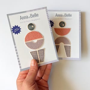 Könnte beinhalten: Zwei Bügelflicken mit geometrischen Designs. Jeder Flicken hat einen kreisförmigen oberen Teil, einen Halbkreis und gestreifte Elemente. Die Flicken befinden sich auf Karten mit dem Text "Anna et Belle" und "Iron-On Patch / Bügelpatch". Der Text "make it your style!" ist ebenfalls sichtbar.