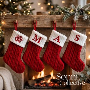 May include: Four red cable-knit Christmas stockings hanging in a row. Each stocking features a white ribbed cuff. The first cuff displays a red embroidered snowflake, while the others feature the red embroidered letters M, A, and S respectively.