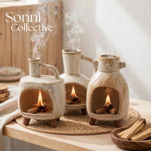 May include: Three cream-colored ceramic incense burners with a speckled finish, each featuring a hollow arched opening, a handle, and three small feet. Each burner contains a burning piece of palo santo wood emitting thin wisps of white smoke.