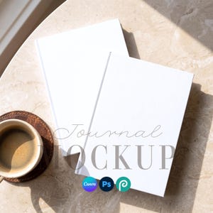 May include: Two blank white journals stacked on top of each other. The text "Journal MOCKUP" is overlaid in the center. A ceramic cup filled with coffee sits on a dark brown circular coaster. The journals have a clean, solid white cover.