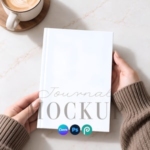 Könnte beinhalten: Ein leeres weißes Journal mit festem Einband, das in den Händen gehalten wird. Der Einband zeigt den Text Journal MOCKUP in einer Kombination aus Schreib- und Serifenschrift sowie kleine runde Symbole für Canva, Photoshop und ein stilisiertes P.