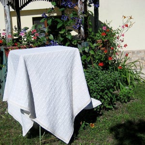 Pu&ograve; includere: Una tovaglia bianca con un motivo a rombi e un bordo in pizzo. La tovaglia &egrave; drappeggiata su un tavolo e posizionata su uno sfondo di piante verdi e fiori.