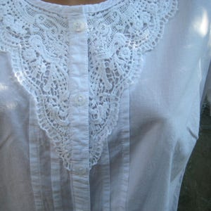 May include: White button-down shirt with a detailed, openwork lace collar. The collar is a prominent feature, with intricate floral and geometric patterns. The shirt has a classic, tailored design with vertical pleats and visible buttons.