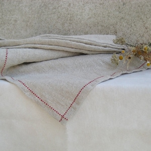 May include: A natural linen tablecloth with a red stitched border. The tablecloth is folded in half and draped over a white surface.