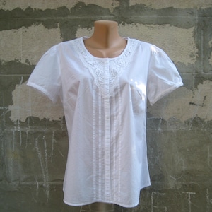 May include: A white short-sleeved blouse with a round neckline and a button-down front. The blouse features delicate lace detailing around the neckline and sleeve edges. Vertical pleats run down the front of the blouse.