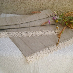 May include: A beige linen table runner with a delicate white lace trim. The runner is folded in half and has a subtle striped pattern.