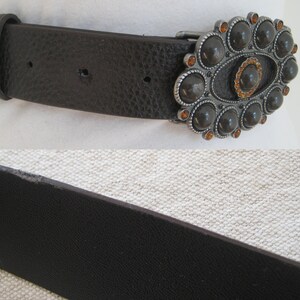 May include: A brown leather belt with a large, ornate silver buckle. The buckle is decorated with black and orange gemstones.