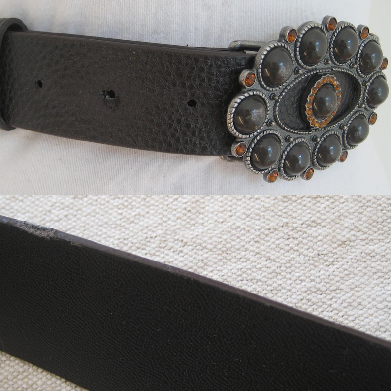 May include: A brown leather belt with a large, ornate silver buckle. The buckle is decorated with black and orange gemstones.