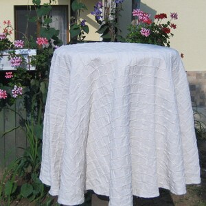 Pu&ograve; includere: Un tovaglia rotonda bianca con un design testurizzato trapuntato. La tovaglia &egrave; drappeggiata su un tavolo e ha un bordo smerlato.