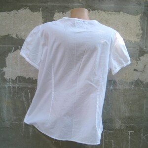 May include: A white short-sleeved blouse with a V-neck and delicate lace trim on the sleeves. The back of the blouse is visible, showing vertical seams and a slightly gathered design. The fabric appears to be lightweight and slightly textured.