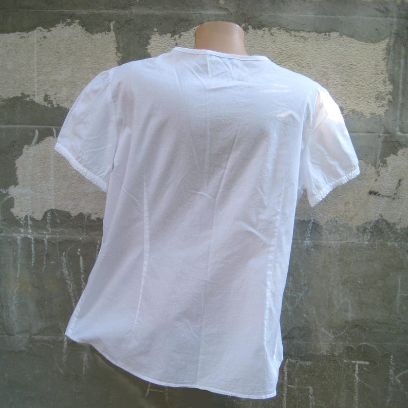 May include: A white short-sleeved blouse with a V-neck and delicate lace trim on the sleeves. The back of the blouse is visible, showing vertical seams and a slightly gathered design. The fabric appears to be lightweight and slightly textured.