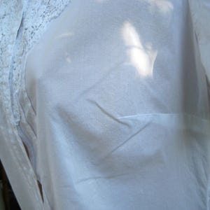 May include: A white, semi-sheer blouse with a lace trim detail on the shoulder. The fabric appears to be lightweight and slightly wrinkled. The blouse has a simple, classic design.