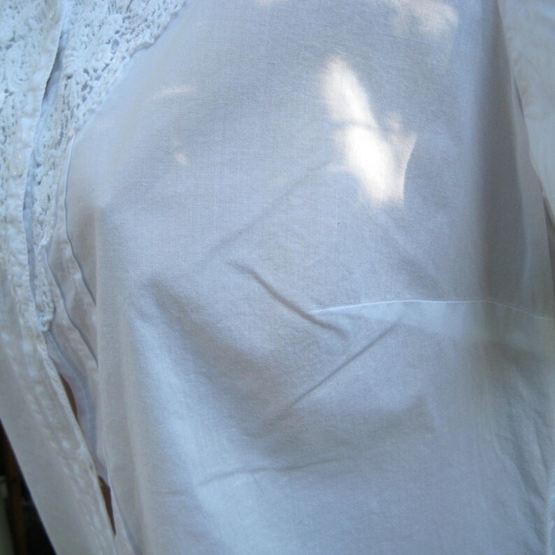 May include: A white, semi-sheer blouse with a lace trim detail on the shoulder. The fabric appears to be lightweight and slightly wrinkled. The blouse has a simple, classic design.