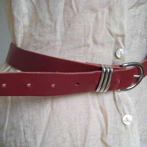 May include: A red leather belt with a silver-colored buckle and loops. The belt is worn over a cream-colored, button-down shirt. The belt has several holes for adjustment.